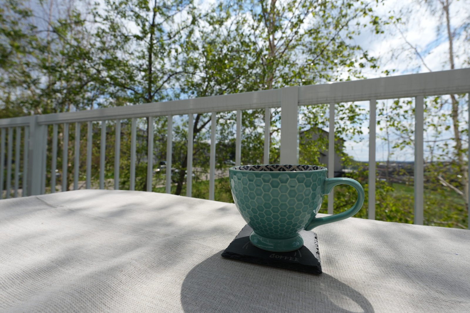 Outdoor balcony lifestyle photo featuring a turquoise coffee mug resting on a personalized engraved slate coaster on a linen-covered table with bright natural sunlight and green trees in the background.