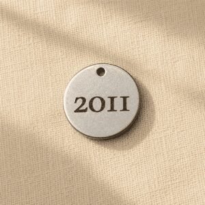 Personalized silver-tone earring charm engraved with a custom year, displayed on soft beige linen fabric in warm natural light. Memorial jewelry detail for pet remembrance earrings.