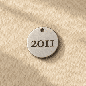Personalized silver-tone earring charm engraved with a custom year, displayed on soft beige linen fabric in warm natural light. Memorial jewelry detail for pet remembrance earrings.