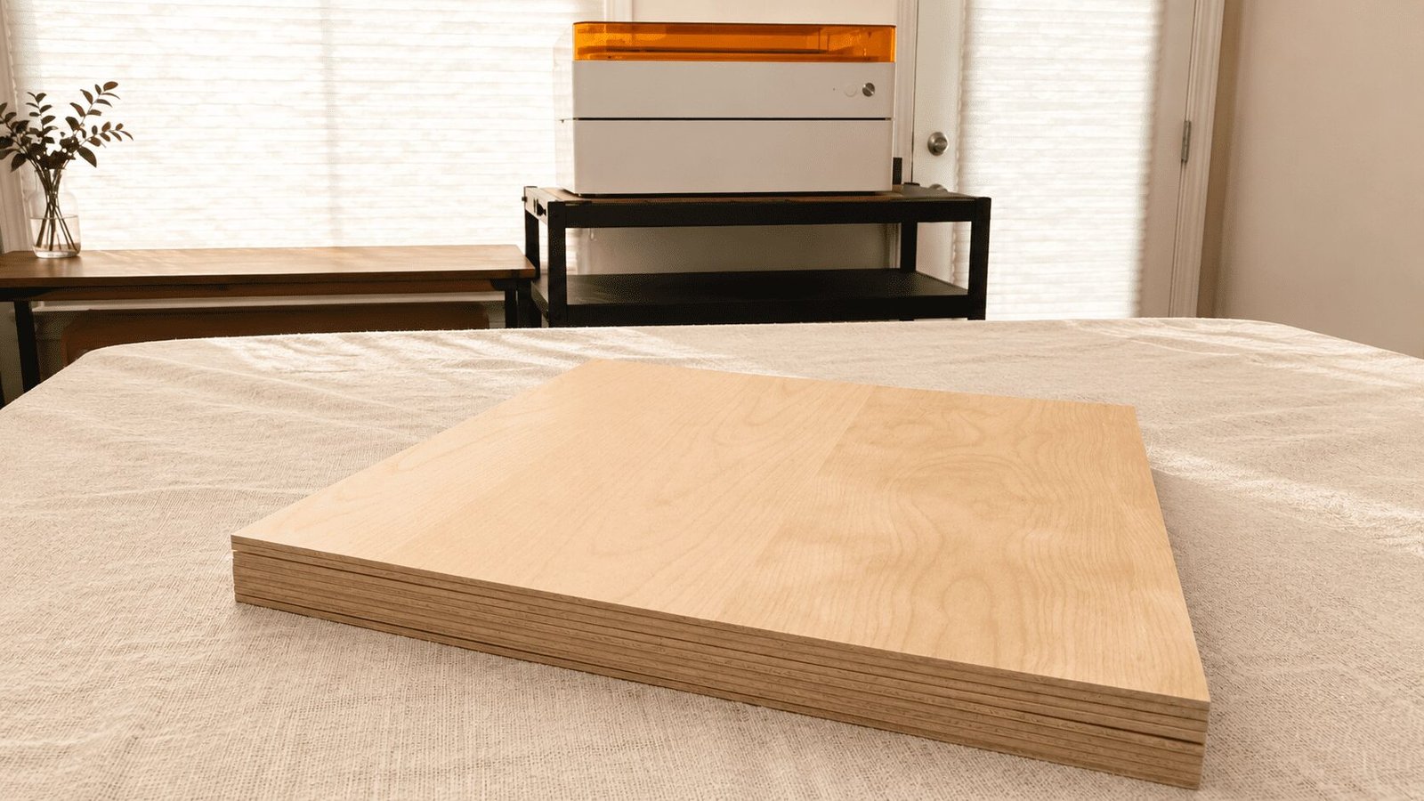 Stacked plywood sheets placed on a fabric-covered worktable with a laser cutting machine in the background.