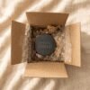 Top-down view of a black round slate memorial coaster engraved with “Forever Loved,” carefully packaged inside a kraft cardboard shipping box with protective bubble wrap on cream linen fabric.