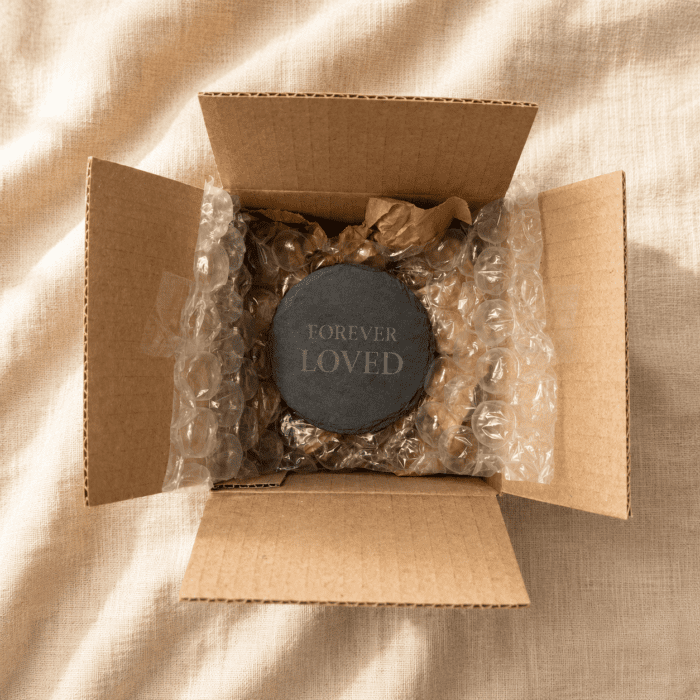 Top-down view of a black round slate memorial coaster engraved with “Forever Loved,” carefully packaged inside a kraft cardboard shipping box with protective bubble wrap on cream linen fabric.