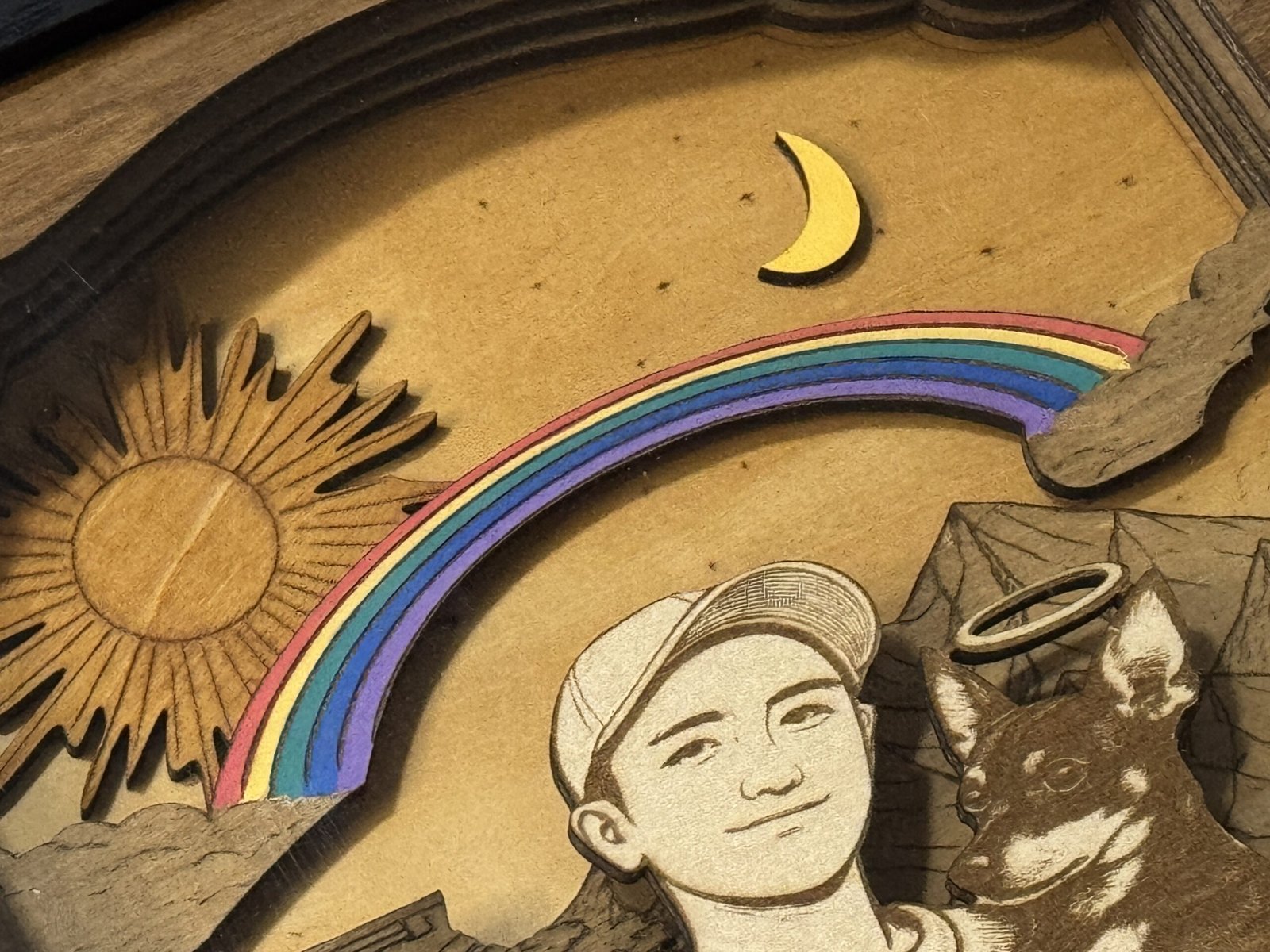 Close-up detail of a handcrafted wooden pet memorial shadow box showing a layered rainbow bridge, engraved sun and moon, and custom portrait engraving of a pet and owner.
