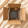 Personalized engraved slate pet memorial coaster packaged securely inside a kraft shipping box with protective bubble wrap, displayed on soft cream linen fabric in warm natural lighting.