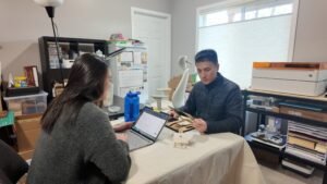 Two people working together in a small home studio with a laptop, handcrafted products, and a laser cutting machine.