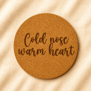Round cork coaster featuring the engraved quote “Cold nose warm heart” in handwritten script typography on a warm cream linen background.