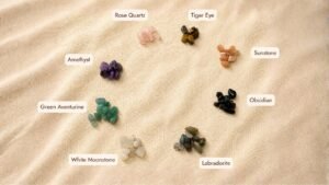 Flat lay photograph displaying labeled crystal chip color options on soft beige linen fabric, including Rose Quartz, Tiger Eye, Sunstone, Obsidian, Labradorite, White Moonstone, Green Aventurine, and Amethyst gemstones.