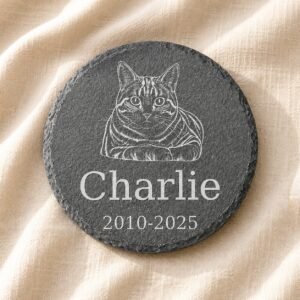 Personalized round pet memorial slate coaster engraved with a detailed cat portrait illustration, pet name Charlie, and memorial dates on soft cream linen fabric.