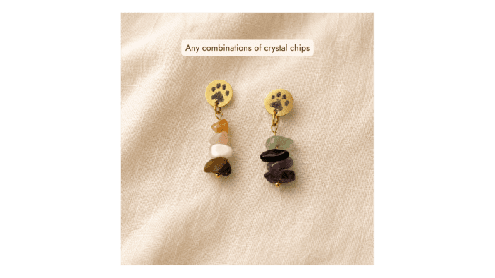Two customizable paw print gemstone earrings displayed on soft cream linen fabric, showing different crystal chip combinations in warm gold-tone settings.
