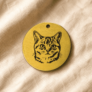Round gold engraved cat portrait keychain displayed on soft beige linen fabric with warm natural lighting.