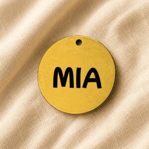 Round gold personalized pet keychain with the engraved name “MIA” displayed on soft beige linen fabric background.
