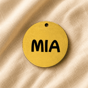 Round gold personalized pet keychain with the engraved name “MIA” displayed on soft beige linen fabric background.