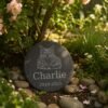 Personalized round pet memorial garden slate engraved with a cat portrait, pet name Charlie, and memorial dates, displayed among flowers, stones, and greenery in a peaceful garden setting.