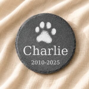 Round engraved pet memorial slate coaster with white paw print, pet name Charlie, and memorial dates on soft cream linen fabric.