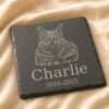 Close-up product photograph of a square engraved pet memorial slate coaster featuring a detailed cat portrait, the name Charlie, and memorial dates on soft cream linen fabric.