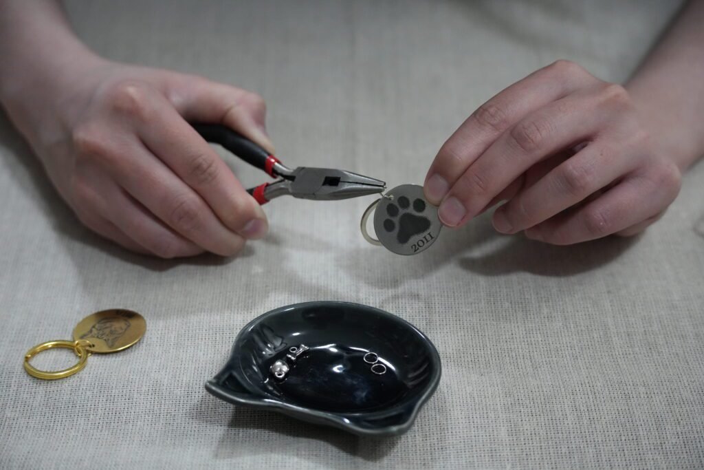 Hands assembling a personalized pet memorial keychain using jewelry pliers, with engraved paw print charm and hardware components on a neutral linen workspace.