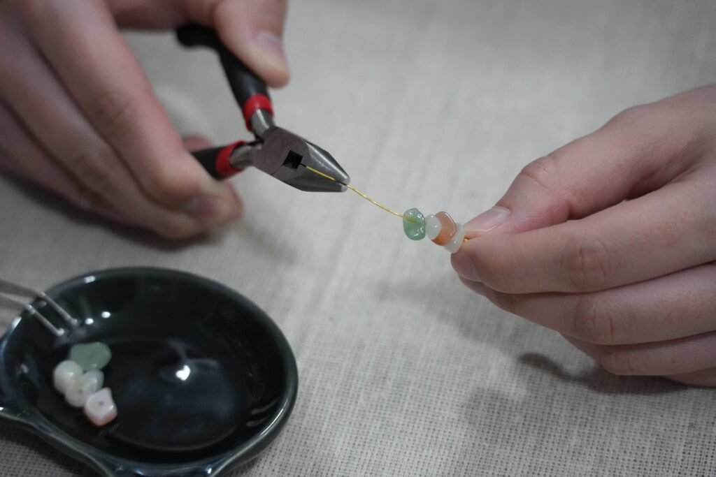 Close-up behind-the-scenes photo of handmade gemstone earrings being assembled with jewelry pliers and natural crystal beads on a neutral linen workspace.