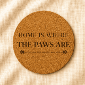 Round cork coaster featuring the engraved quote “Home Is Where The Paws Are” with decorative paw print divider accents on a warm cream linen background.