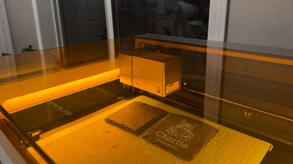 Behind-the-scenes photograph of a laser engraving machine engraving a personalized pet memorial slate coaster with a custom cat portrait and memorial text inside a warm-lit workshop.