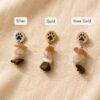 Flat lay photo showing three paw print gemstone earrings in silver, gold, and rose gold finishes on soft cream linen fabric with labeled color options.