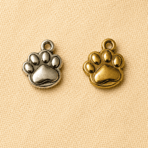 Silver and gold paw print memorial charm pendants displayed on a beige linen background, designed for pet remembrance jewelry, keepsakes, and personalized memorial gifts.