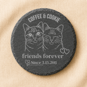 Round black slate coaster engraved with custom portraits of two cats, Coffee and Cookie, featuring the phrase “Friends Forever” and a personalized date, displayed on a warm cream linen background.