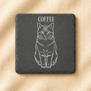 Square black slate coaster with an engraved portrait of an orange tabby cat named Coffee on a warm cream linen background.