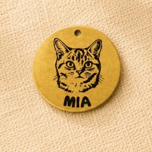 Custom engraved gold pet memorial keychain charm featuring a cat portrait and pet name on a round gold-colored metal tag displayed on beige linen fabric.