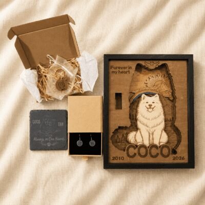 Flat lay of personalized pet memorial keepsakes including a wooden rainbow bridge shadow box, engraved pet keychain in gift packaging, memorial slate coaster, and custom pet earrings arranged on warm neutral linen fabric.
