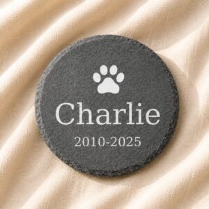 Round engraved pet memorial slate stone with paw print icon, pet name Charlie, and memorial dates displayed on soft cream linen fabric.