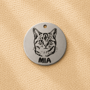 Custom engraved silver pet memorial keychain charm featuring a cat portrait and pet name on a round silver-colored metal tag displayed on beige linen fabric.
