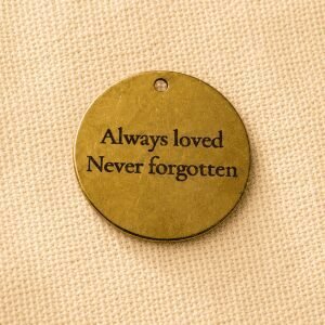 Round gold pet memorial keychain charm engraved with the message “Always loved, Never forgotten” displayed on a beige linen fabric background.
