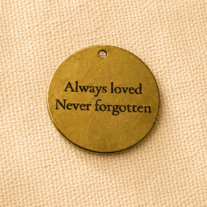 Round gold pet memorial keychain charm engraved with the message “Always loved, Never forgotten” displayed on a beige linen fabric background.