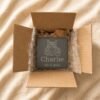 Top-down packaging photograph of a personalized pet memorial slate coaster engraved with a cat portrait, securely packed inside a cardboard shipping box with protective bubble wrap and kraft packing paper on neutral linen fabric.