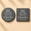 Comparison photograph of round and square personalized pet memorial slate coasters engraved with a detailed cat portrait, pet name Charlie, and memorial dates on neutral woven fabric.