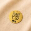 Close-up photograph of a personalized gold-tone pet charm engraved with a cat portrait and the name “MIA,” displayed on soft cream linen fabric.