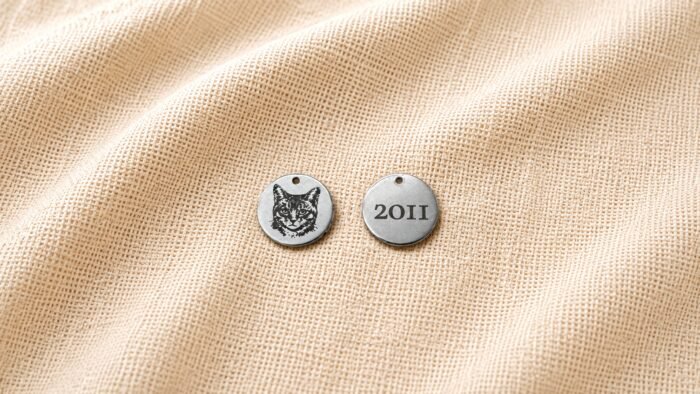 Front and back view of a personalized engraved pet charm on beige linen fabric, featuring a custom cat portrait on one side and the year “2011” engraved on the reverse side.