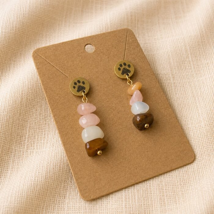 Paw Print Gemstone Earrings Close Up Product Photo Close-up product photograph of handmade paw print gemstone earrings displayed on a kraft jewelry card over soft beige linen fabric, featuring warm earthy gemstone tones and gold accents.